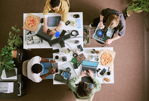 Overhead view of a diverse team collaborating on computer hardware repairs and software coding at a cluttered tech workstation with laptops and pizza. 