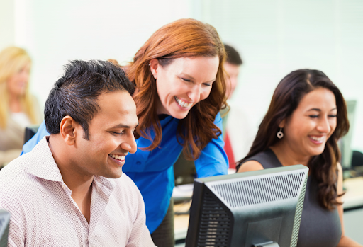 A smiling female instructor or manager leans over a desk to assist a male colleague at a computer, with other diverse professionals working in a bright, modern office background.