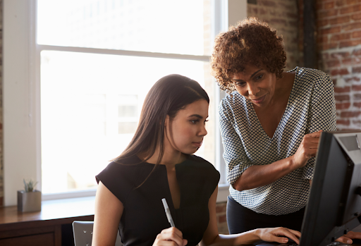 A senior female professional with curly hair mentoring a younger colleague at a computer desk in an office with exposed brick walls. 