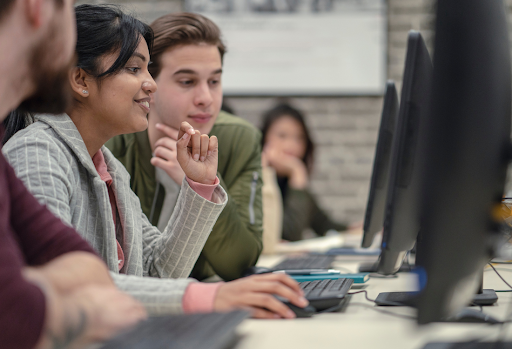 A diverse group of young students in a classroom setting, focused intently on a computer screen while discussing a technical project. 