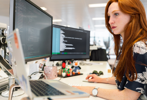 A focused young redhead woman with freckles coding at a dual-monitor workstation in a professional office environment. 
